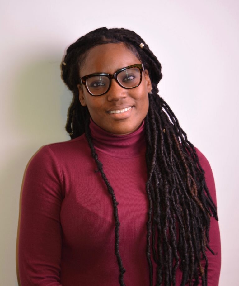Professional headshot of a woman with glasses and long braids wearing a burgundy turtleneck against a neutral background